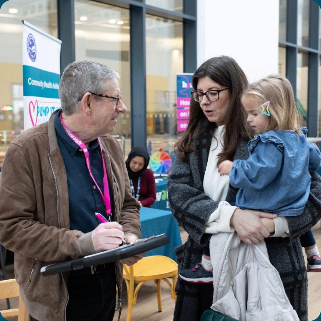 An image of a male Healthwatch Engagement Officer listening to a mother as she is holding her daughter. The mother is talking to the Engagement Officer. The mother is sharing some of her health and social care experiences. They are at a public event and they are standing in front of a Healthwatch table.